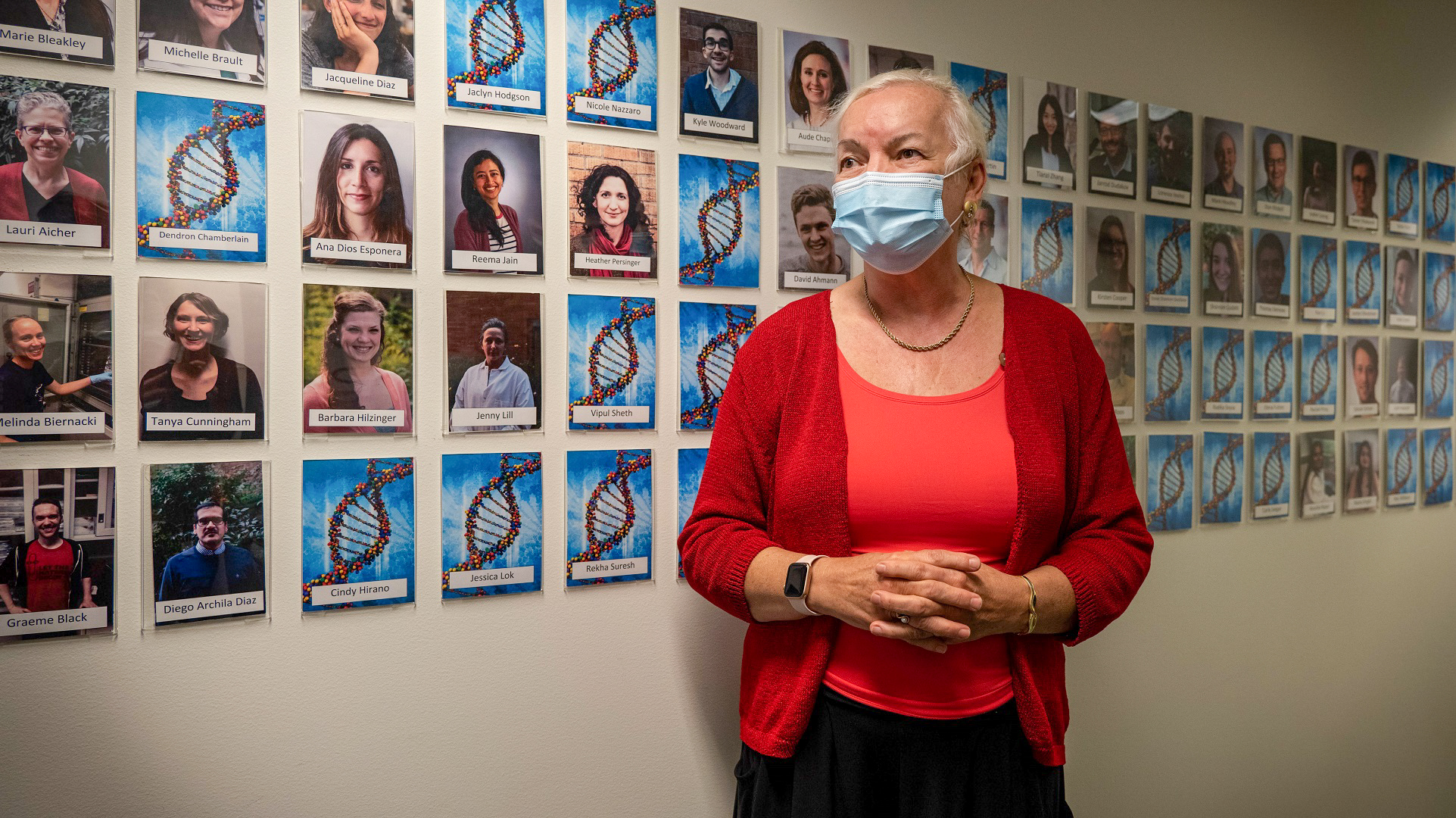 Natalie Duerkopp, a woman with cropped white hair and a vibrant red cardigan, standing in front of a wall of Fred Hutch lab staff.